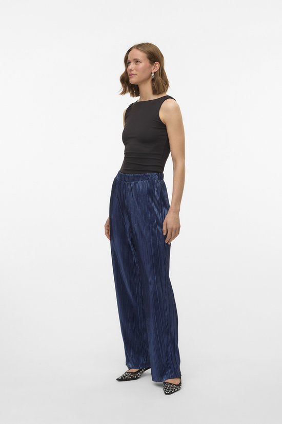 Model draagt een donkerblauwe, glanzende, geplooide broek met wijde pijpen.