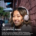 Vrouw in lichtgrijze koptelefoon bij bakstenen muur, met tekst 'INGEBOUWDE LAB SPATIAL AUDIO'.