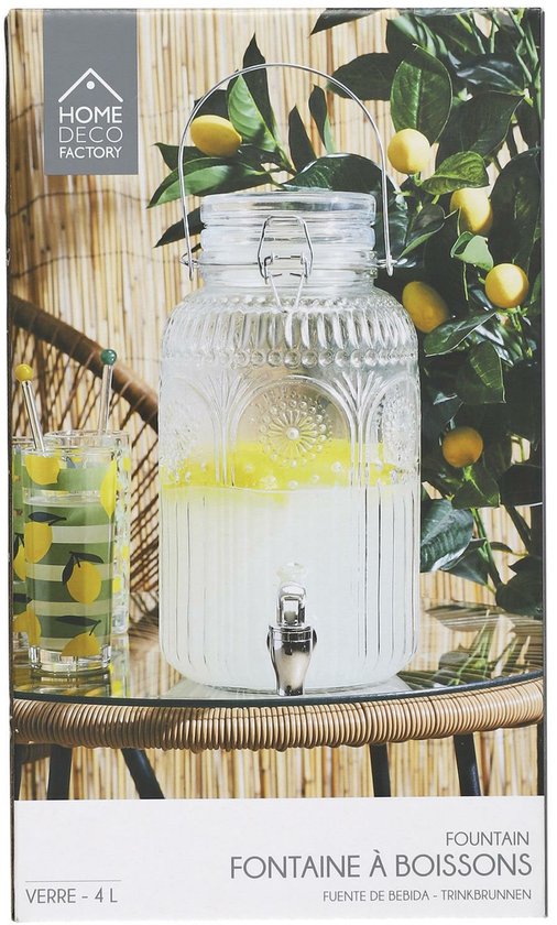 Glazen drankdispenser met kraantje, gevuld met gele drank, naast glazen met citroenprint. Staat op een tafel onder een citroenboom.