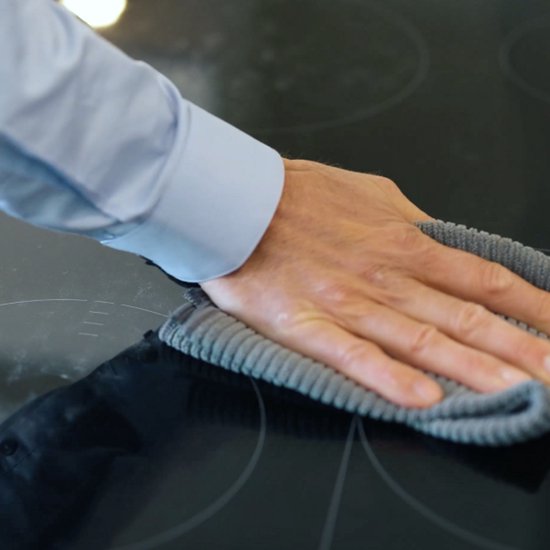 Une main avec un chiffon en microfibre gris nettoie une plaque de cuisson en verre.