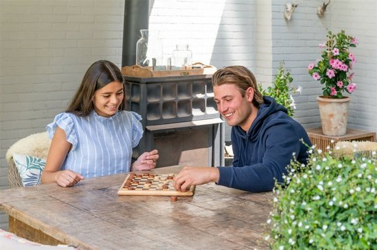 Een jong stel lacht terwijl ze buiten op een houten tafel een damspel spelen.