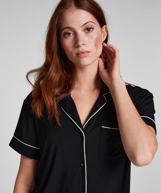 Model met rood haar draagt een zwart pyjamashirt met korte mouwen en witte piping. Het shirt heeft een open kraag en borstzakje.
