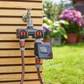 GARDENA besproeiingscomputer met kraan en slang, op houten wand. Groene tuin met bloemen.