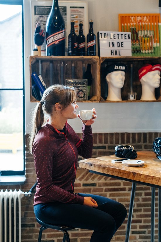 Vrouw in een bordeauxrode longsleeve fietstop met 'love' patroon, drinkt koffie aan een tafel.