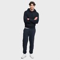 Man met gekruiste armen, draagt donkerblauwe hoodie met capuchon, kangoeroezak en een klein logo. Draagt een witte T-shirt en navy joggingbroek.