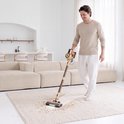Man in beige trui en witte broek zuigt een vloerkleed met gouden en bruine steelstofzuiger.