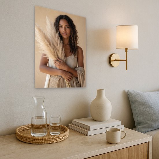 Canvas schilderij van een vrouw met pampasgras boven een houten dressoir met glazen karaffen en een schemerlamp.