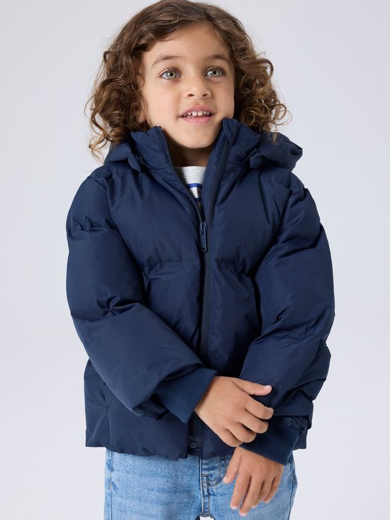 Jongen met krullend haar draagt een donkerblauwe pufferjas met capuchon, gestreept grijs-wit shirt en spijkerbroek.