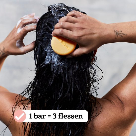 Vrouw wast haar zwarte haar met een oranje shampoo bar. Tekst: '1 bar = 3 flessen'.
