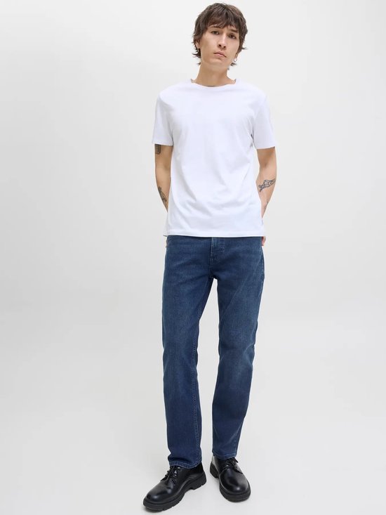 Man met tatoeages draagt een witte T-shirt en een donkerblauwe jeans met rechte pijpen.