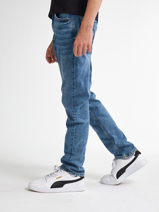 Model draagt een blauwe Petrol Industries jeans met regular tapered fit, gecombineerd met witte Puma sneakers.