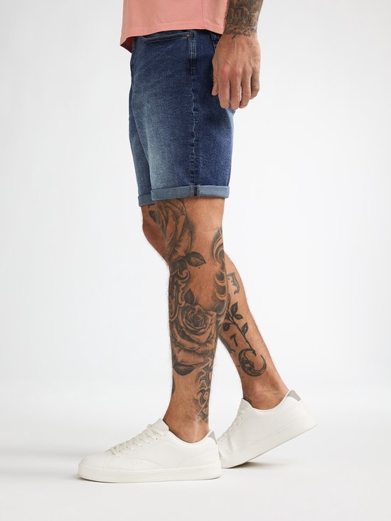Model draagt een Petrol Industries regular-fit jeansshort in denimblauw met opgerolde broekspijpen en witte sneakers.