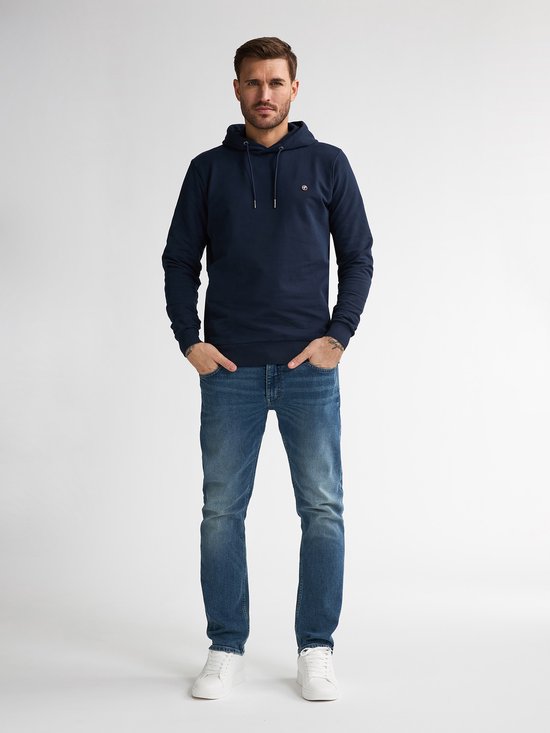 Model draagt een marineblauwe hoodie met logo en een straight fit spijkerbroek in vintage blauw met witte sneakers.
