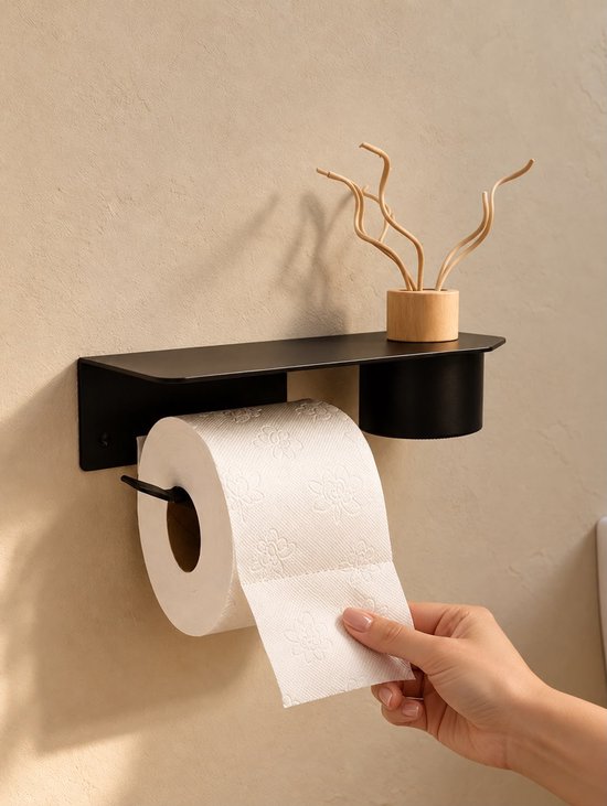 Zwarte toiletrolhouder aan de muur met plank, houten diffuser en hand die toiletpapier vasthoudt.
