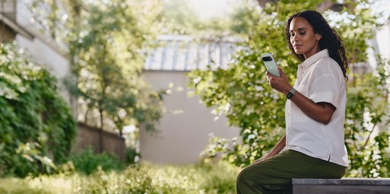 Vrouw in wit overhemd en olijfgroene broek kijkt op haar smartphone met een olijfgroene Fitbit Edge om haar pols.