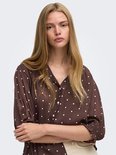 Model draagt bruine blouse met witte polkadotprint, V-hals en knopen.