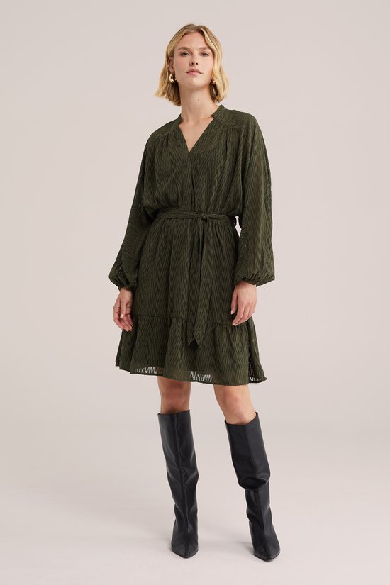 Dames jurk in donkergroen met een strikceintuur, lange mouwen en een gelaagde rok. Gekleed met zwarte laarzen.