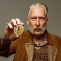 Oudere man met grijze snor en tatoeages, die een blikje Reuzel baardbalsem vasthoudt.