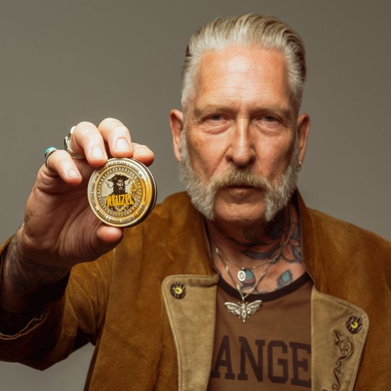 Oudere man met grijze snor en tatoeages, die een blikje Reuzel baardbalsem vasthoudt.