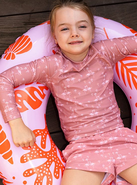 Een meisje in een roze zwemshirt met flamingo's, liggend in een roze zwemband met koraal- en schelpmotief.