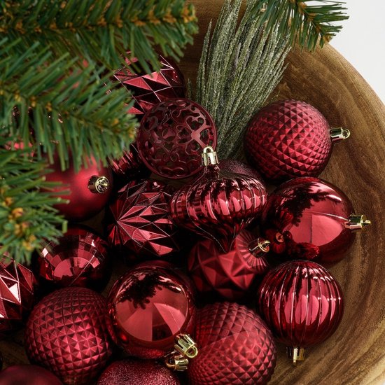 Donkerrode kerstballen met diverse texturen, waaronder glad, geribbeld en facet, in een houten kom met dennentakken.