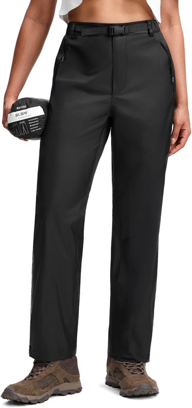 Femme portant un pantalon de pluie noir avec ceinture, tenant un pantalon de pluie enroulé. Arrière-plan de studio vide.