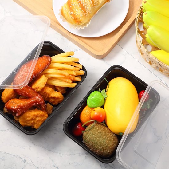 Twee zwarte voedselcontainers, één met kip en frietjes, de andere met fruit en groenten, naast bakkerijproducten en bananen.