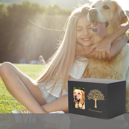 Meisje omhelst haar hond naast een zwarte houten urn met goudkleurige levensboom en tekst 'Forever in our heart'.