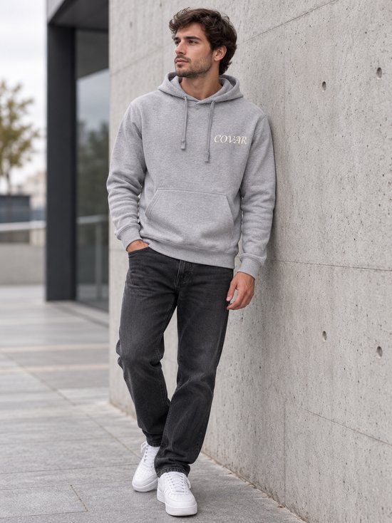 Man leunt tegen muur, draagt grijze hoodie met 'COVAR' print.