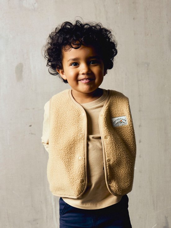 Kind met krullend haar draagt een beige teddy-gilet over een bijpassend shirt.