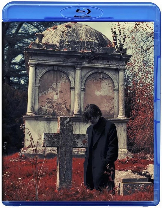 Jonge man in zwarte jas bij een kruis en mausoleum in een verlaten, roodgetinte begraafplaats.