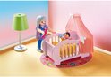 Een Playmobil oma staat bij een roze babywieg met hemel en een groene lamp, op een rond vloerkleed.
