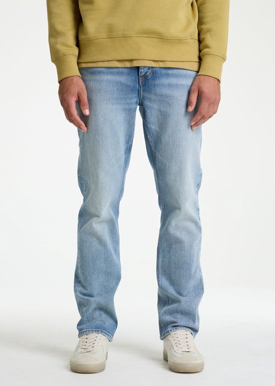 Man met lichtblauwe spijkerbroek en okergele trui, recht van voren gezien.