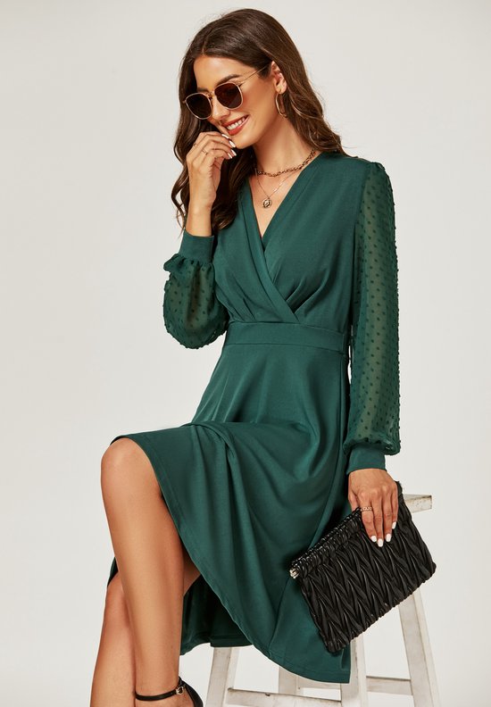 Model zittend op een kruk, draagt een donkergroene midi-jurk met wikkelkraag en pofmouwen met stippen. Accessoires: zonnebril, ketting, tas, hoge hakken.