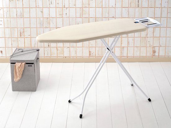 Crèmekleurige strijkplank uitgeklapt met wit metalen frame, naast een grijze wasmand.