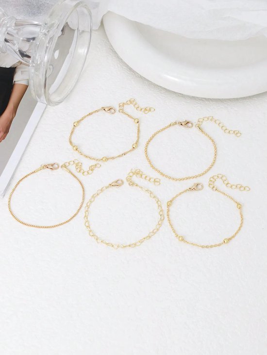 Vijf gouden armbandjes met diverse schakels en gouden kraaltjes, met karabijnsluiting, los op een witte ondergrond.