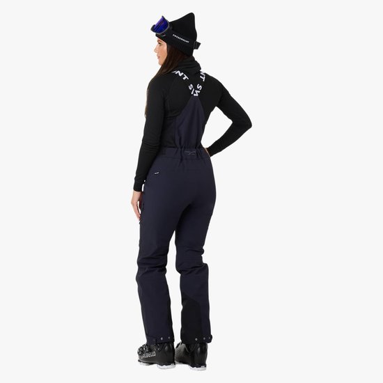 Vrouw met donkerblauwe skibroek met bretels, zwart coltrui, ski-laarzen en skibril, van achteren gezien.