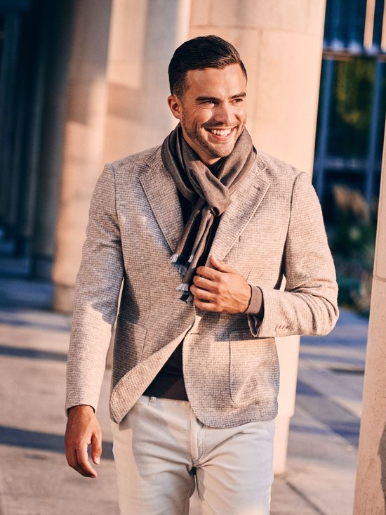 Man met grijze sjaal en beige colbert met een donkerbruine trui en beige broek.
