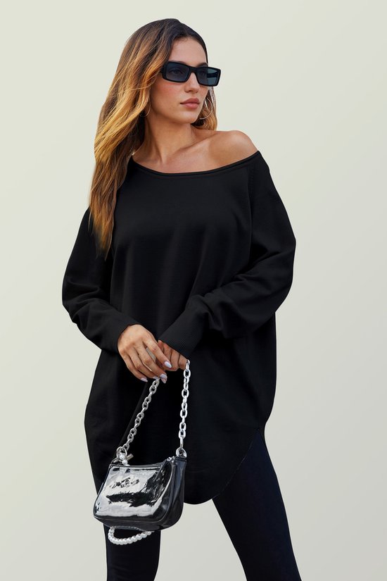 Model draagt een oversized zwarte gebreide trui met een off-shoulder halslijn, met een zwarte tas met zilveren ketting.