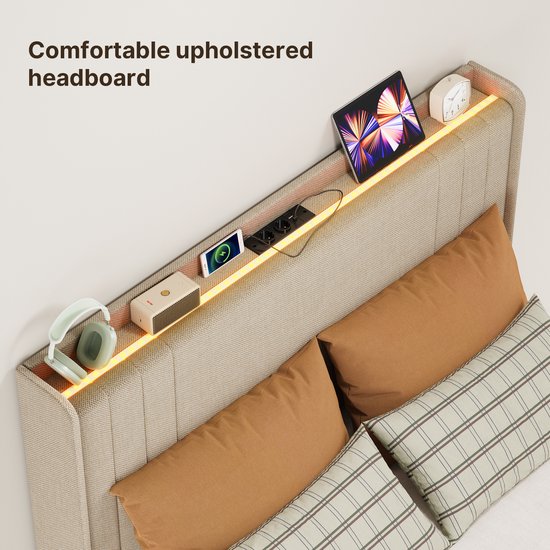 Een detail van het beige gestoffeerde hoofdbord met opbergruimte, verlichting, tablet, telefoon en koptelefoon.
