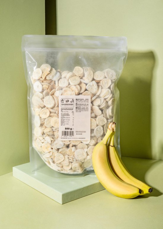 Een doorzichtige zak met gevriesdroogde bananenschijfjes. Twee bananen liggen ernaast op een lichtgroen platform.