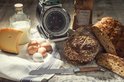 Verschillende broden, kaas, eieren, melk en een broodmes met olijfhouten handvat op een houten ondergrond.