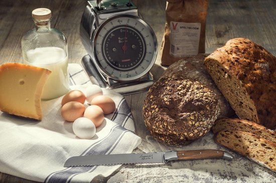 Verschillende broden, kaas, eieren, melk en een broodmes met olijfhouten handvat op een houten ondergrond.