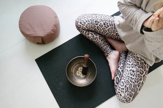 Vrouw in luipaardprint legging mediteert naast een taupe meditatiekussen en klankschaal.