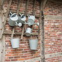 Vier zinken gieters hangen aan een houten rek. Twee zinken emmers hangen lager aan het rek.