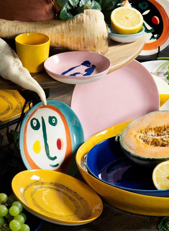 Een serveerschaal met een kindertekening van een gezicht, met blauwe rand, oranje haar en gele en rode wangen, omringd door fruit.