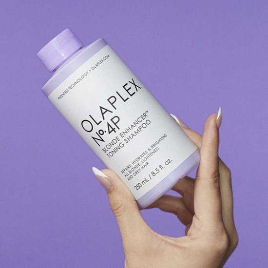 Hand houdt een fles Olaplex No.4P Blonde Enhancer Toning Shampoo vast tegen een paarse achtergrond.