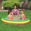 Een vrouw kijkt lachend toe hoe een jongen en een meisje spelen in een driekleurig opblaasbaar zwembad in de tuin.
