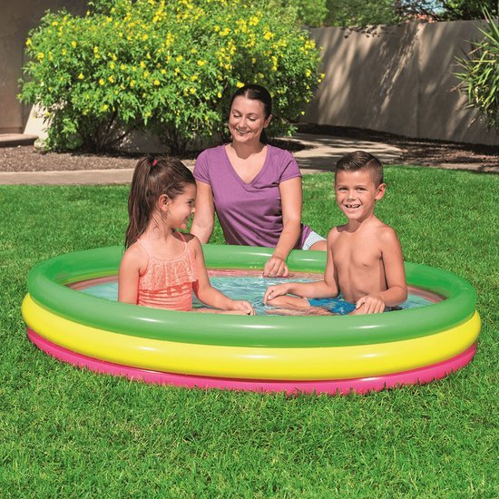 Een vrouw kijkt lachend toe hoe een jongen en een meisje spelen in een driekleurig opblaasbaar zwembad in de tuin.