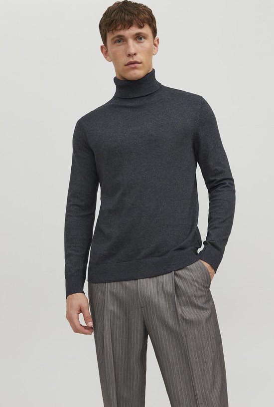 Man staand, draagt een donkergrijze coltrui en een grijze pantalon met krijtstrepen.